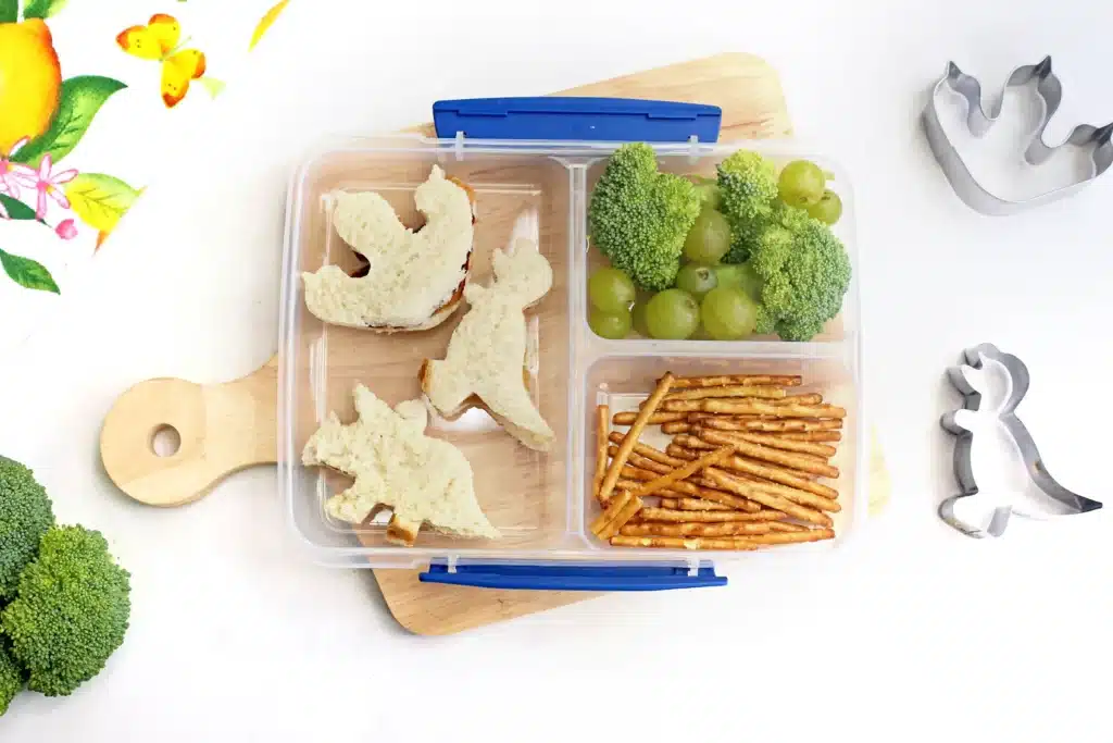 Dinosaur Lunch Idea - Cultivating Brilliant Minds a lunchbox with sandwiches cut into dinosaurs, pretzels, broccoli, and green grapes