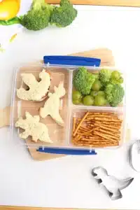 a lunchbox with sandwiches cut into dinosaurs, pretzels, broccoli, and green grapes