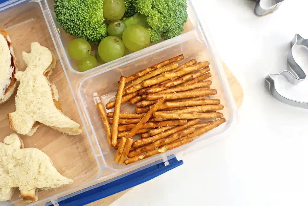 Dinosaur Lunch Idea - Cultivating Brilliant Minds a lunch box idea for kids that has dinosaur sandwiches, broccoli trees, and pretzels in a lunchbox container