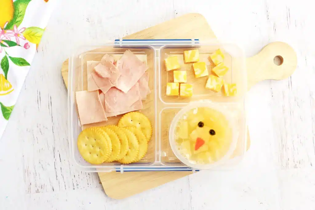 Spring Lunchbox Idea for Kids - Cultivating Brilliant Minds a spring lunchbox idea for kids that has ham, crackers, and cheese in a lunchbox along with a pineapple fruit cup with a chick face drawn on the lid