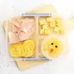 Spring Lunchbox Idea for Kids - Cultivating Brilliant Minds a spring lunchbox idea for kids that has ham, crackers, and cheese in a lunchbox along with a pineapple fruit cup with a chick face drawn on the lid