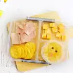 Spring Lunchbox Idea for Kids - Cultivating Brilliant Minds a spring lunchbox idea for kids that has ham, crackers, and cheese in a lunchbox along with a pineapple fruit cup with a chick face drawn on the lide