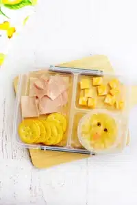 a spring lunchbox idea for kids that has ham, crackers, and cheese in a lunchbox along with a pineapple fruit cup with a chick face drawn on the lide