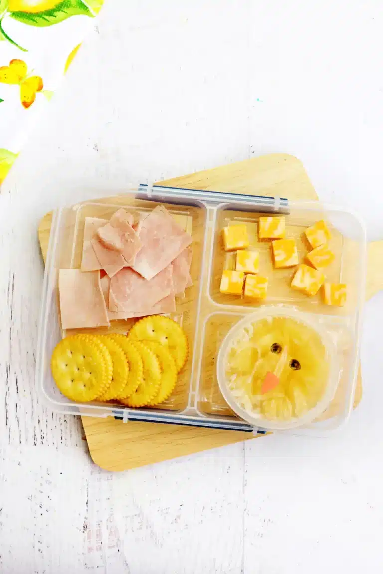 a spring lunchbox idea for kids that has ham, crackers, and cheese in a lunchbox along with a pineapple fruit cup with a chick face drawn on the lide