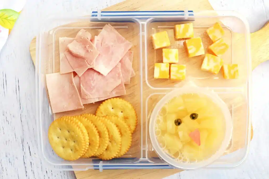 Spring Lunchbox Idea for Kids - Cultivating Brilliant Minds a spring lunchbox idea for kids that has ham, crackers, and cheese in a lunchbox along with a pineapple fruit cup with a chick face drawn on the lid