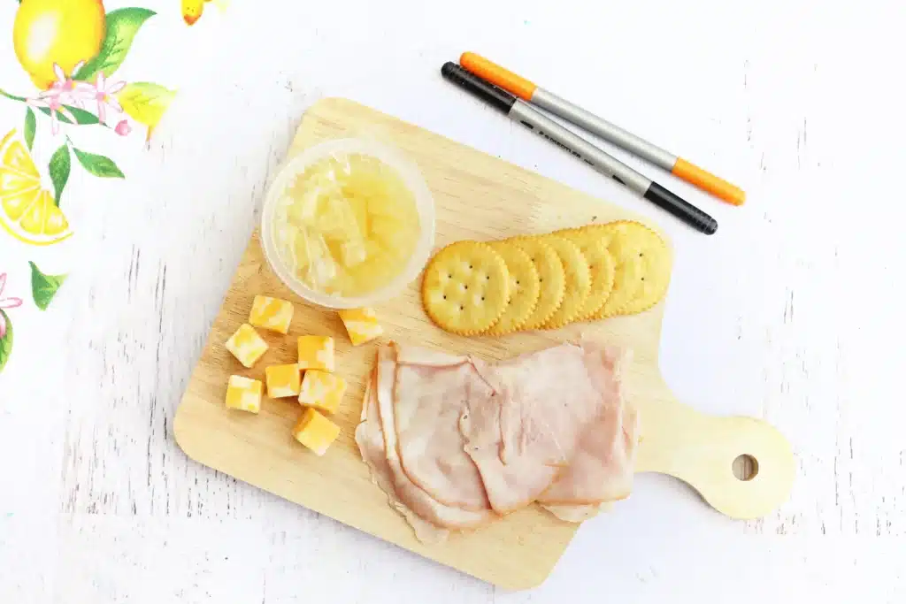 Spring Lunchbox Idea for Kids - Cultivating Brilliant Minds a pineapple fruit cup, ham, crackers, and cubes of cheese on a wooden board