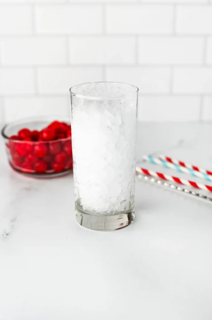 Copycat Sonic Ocean Water - Cultivating Brilliant Minds Tall glass filled with crushed ice on a white countertop, with a blurred bowl of red cherries in the background and red-and-white striped straws nearby.