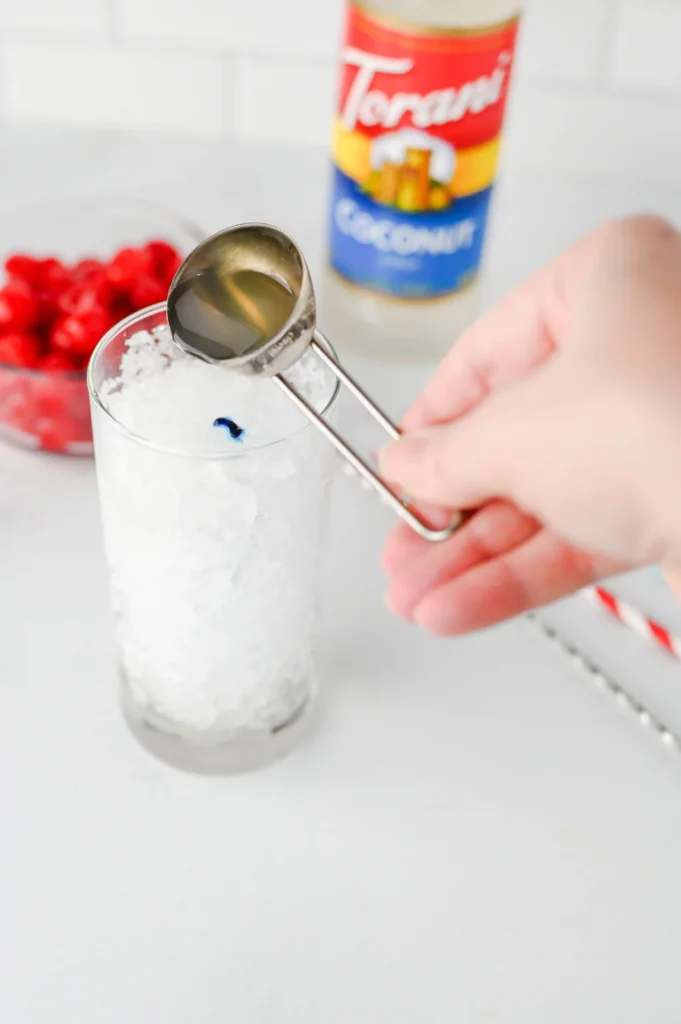 Copycat Sonic Ocean Water - Cultivating Brilliant Minds Person pours coconut syrup from a metal measuring spoon into a tall glass of crushed ice, with a blurred Torani bottle and a bowl of red berries in the background, conveying a drink-prep scene.