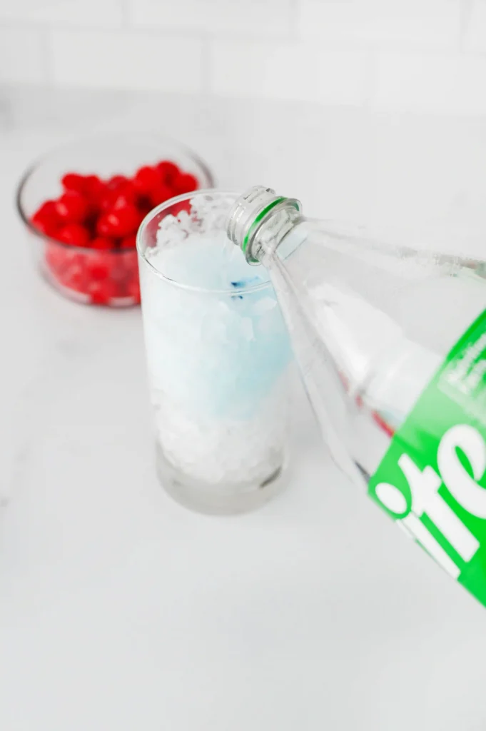 Copycat Sonic Ocean Water - Cultivating Brilliant Minds Sprite is poured from a bottle into a glass filled with ice, with a bowl of red berries blurred in the background.