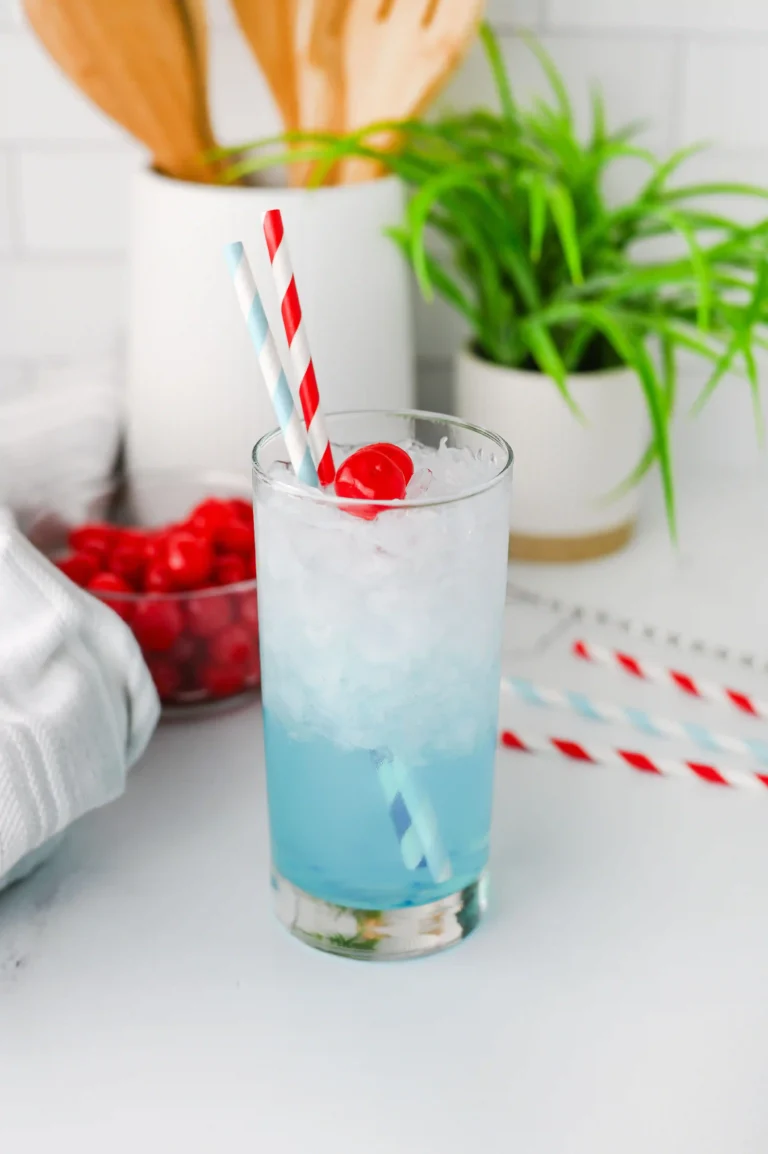 Tall glass of icy blue beverage with red cherries, two striped straws, and a blurred kitchen scene in the background.