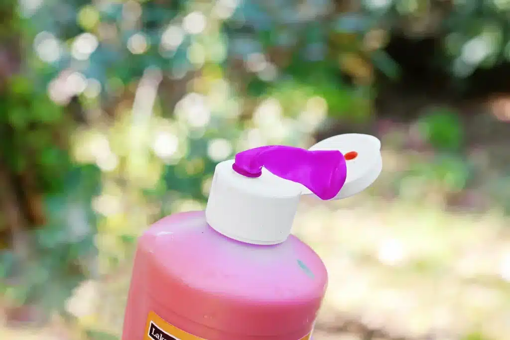 Water Balloon Splatter Art - Cultivating Brilliant Minds Close-up of a pink paint bottle with a white pump dispenser; a purple water balloon rests on top, outdoors with a blurred green garden background.