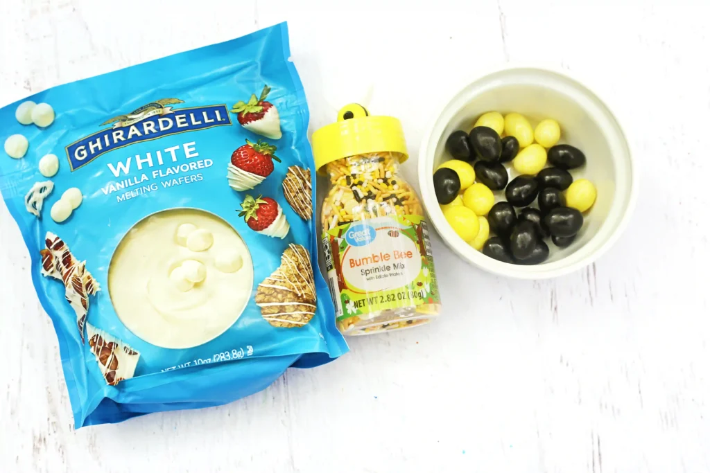Bumble Bee Bark - Cultivating Brilliant Minds Blue bag of Ghirardelli White Vanilla Melting Wafers beside a sprinkles bottle and a bowl of black and yellow jelly beans on a white surface.