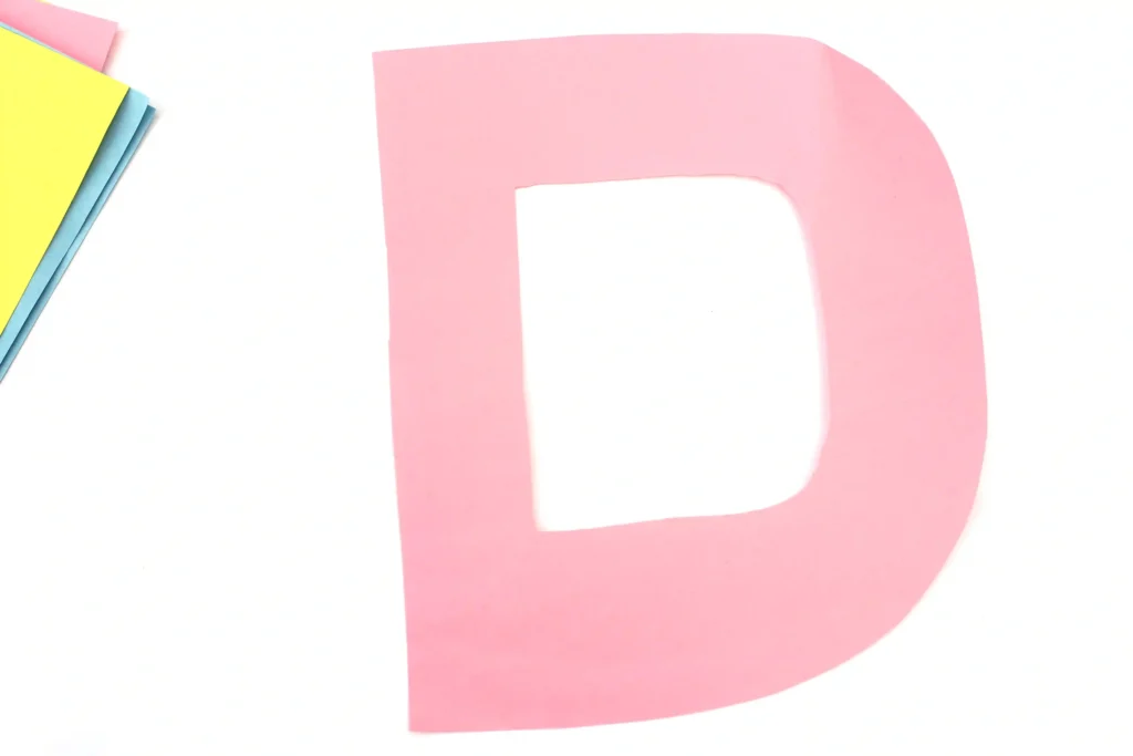 Letter D Craft - Cultivating Brilliant Minds Pink D-shaped paper cutout on a white background with pastel-colored sheets to the left.