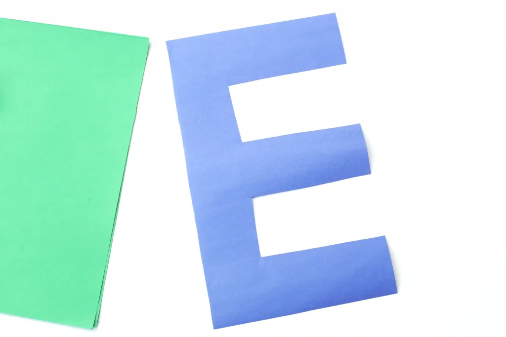 Letter E Earth Craft - Cultivating Brilliant Minds Blue paper cutout in the shape of the letter E on a white background, with a green sheet visible on the left edge.