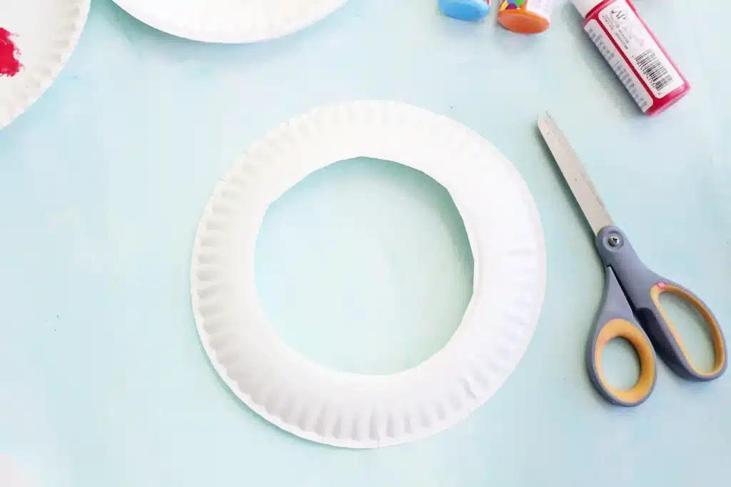 Paper Plate Ring Toss Game - Cultivating Brilliant Minds White paper-plate ring shape on a pale blue craft table with scissors and glue nearby, ready for a kid's craft project