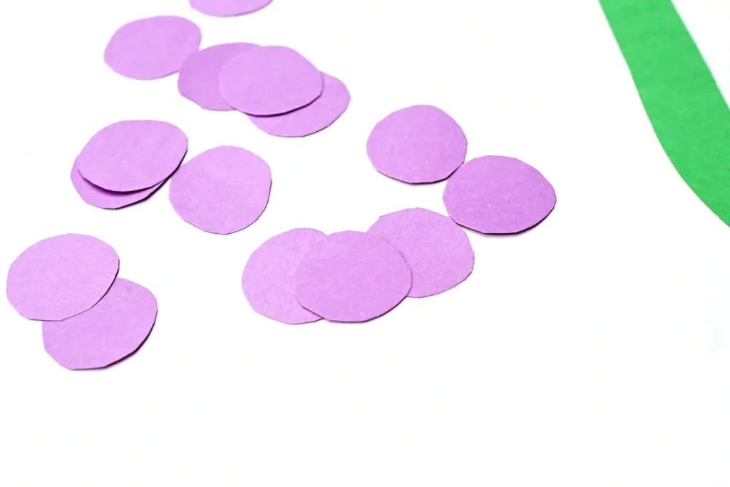 Letter G Grape Craft - Cultivating Brilliant Minds Purple circular purple construction paper pieces scattered on a white surface with a green curved edge in the corner.