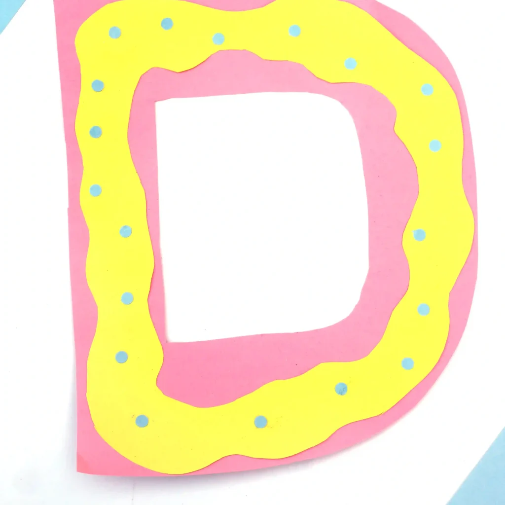 a letter d craft that has a pink letter d with yellow squiggly frosting on top made from yellow construction paper, and blue circles for sprinkles