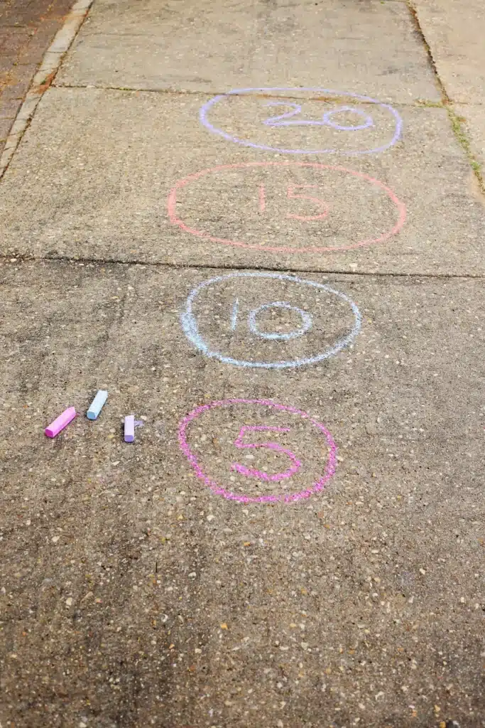 DIY Sponge Toss - Cultivating Brilliant Minds Sidewalk chalk circles with numbers 5, 10, 15, and 20 drawn in pink, blue, red, and purple from bottom to top, with chalk pieces scattered nearby.