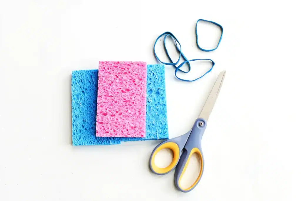 DIY Sponge Toss - Cultivating Brilliant Minds Craft supplies on a white surface: blue and pink sponges with yellow-handled scissors and a blue rubber band nearby.