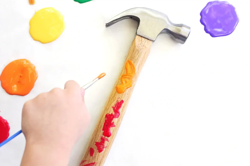 Hammer Father's Day Activity - Cultivating Brilliant Minds Close-up of a hammer on a white surface with bright orange, yellow, and purple paint blobs, while a hand uses a small brush to apply more paint nearby. The hammer will be given as a father's day gift
