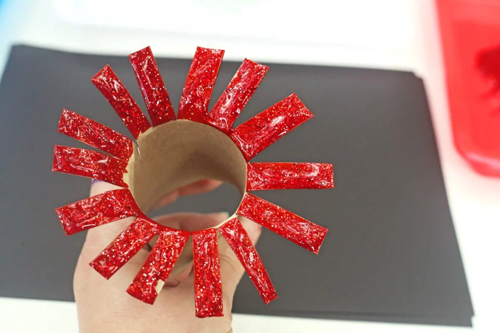 Toilet Paper Roll Firework Craft - Cultivating Brilliant Minds Hand holding a circular toilet paper roll that is cut and covered with red glittery rectangular strips radiating like sun rays.
