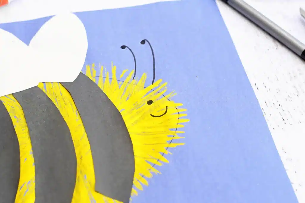 Fork Painted Bee - Cultivating Brilliant Minds Close-up of a handmade paper bee painting on blue background, with yellow/black striped body and a white wing above it.