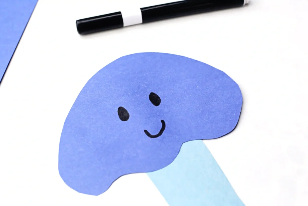 Letter J Jellyfish Craft - Cultivating Brilliant Minds Close-up of a blue paper cutout with a smiling face on a white background, with a marker nearby as craft supplies show through.