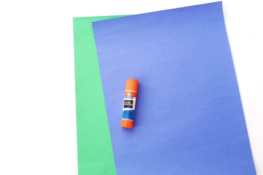Letter E Earth Craft - Cultivating Brilliant Minds Orange Elmer's glue stick lying on blue construction paper overlapped by a green sheet beneath.