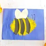 Craft bee on blue card: yellow fluffy body with black stripes and white heart wings, smiling face.