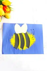 A blue piece of construction paper with a yellow fork painted bee with black construction paper stripes and white wings