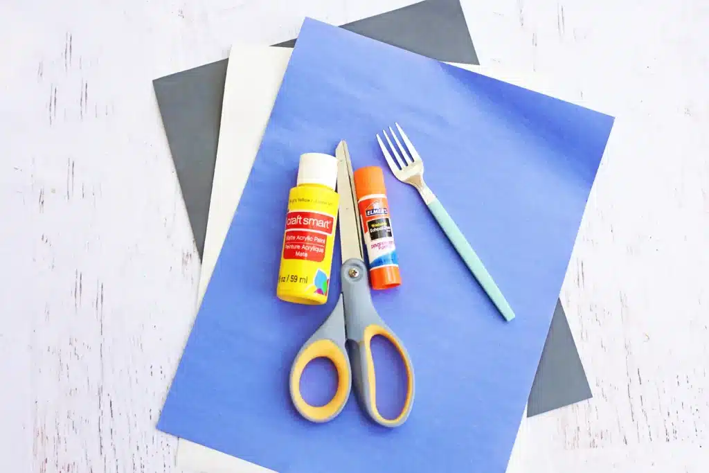 Fork Painted Bee - Cultivating Brilliant Minds Craft supplies arranged on blue paper: scissors, glue bottle, glue stick, a craft knife, and a plastic fork on top of layered color papers.