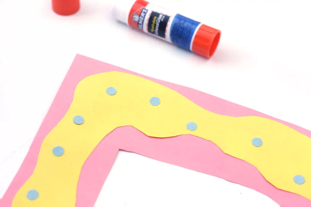 Letter D Craft - Cultivating Brilliant Minds Close-up of a colorful paper craft with pink and yellow shapes and blue dots, with a glue stick in the background.