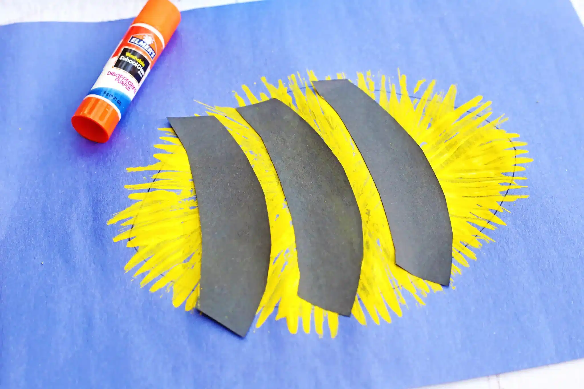 Fork Painted Bee - Cultivating Brilliant Minds Craft project: a blue paper with a yellow sunburst made of paint and three black curved paper strips glued on top to look like a bee, with glue stick nearby.