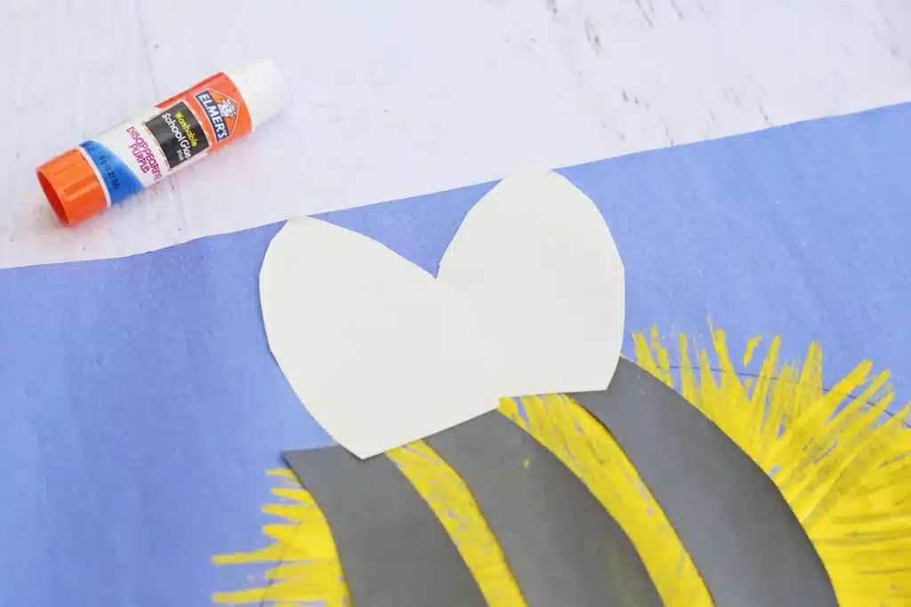 Fork Painted Bee - Cultivating Brilliant Minds White paper wings placed on a blue artwork with yellow sunburst strokes; a glue stick sits in the top-left corner.