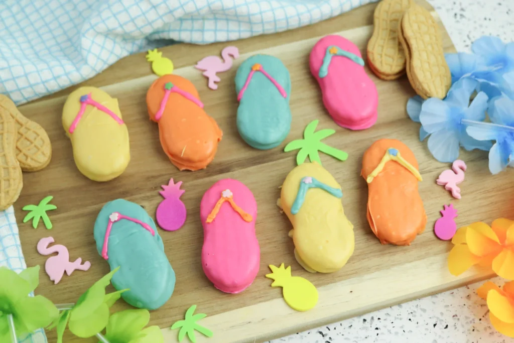 Flip Flop Cookies - Cultivating Brilliant Minds Assorted pastel flip-flop cookies with bright icing on a wooden board, surrounded by tropical shapes and blue flowers (summer cookies).