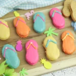 Assorted pastel flip-flop cookies with bright icing on a wooden board, surrounded by tropical shapes and blue flowers (summer cookies).