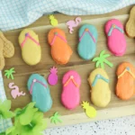 Flip Flop Cookies - Cultivating Brilliant Minds Pastel flip-flop cookies with bright icing on a wooden board, surrounded by tropical paper shapes and a blue checkered towel.