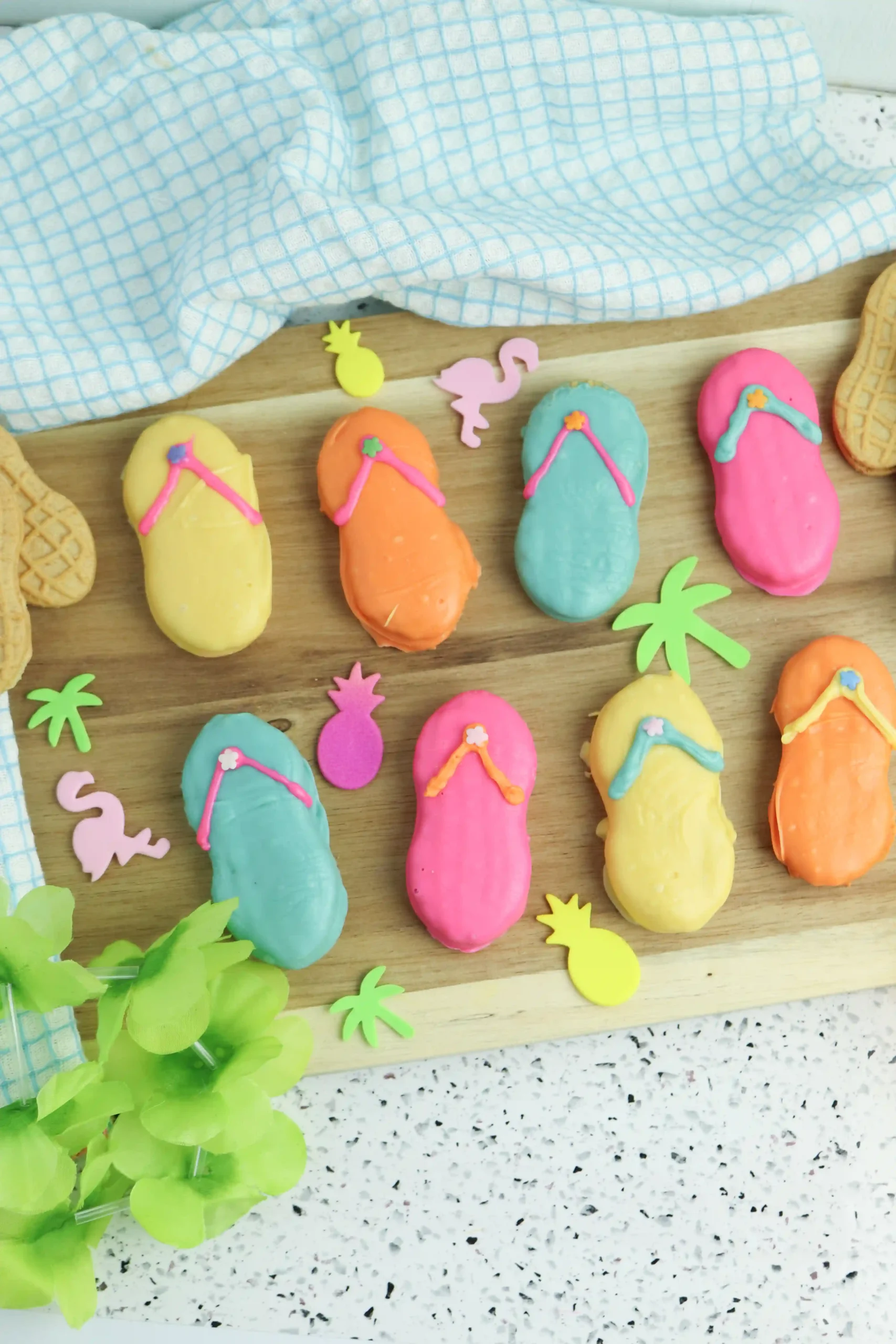 Pastel flip-flop cookies with bright icing on a wooden board, surrounded by tropical paper shapes and a blue checkered towel.