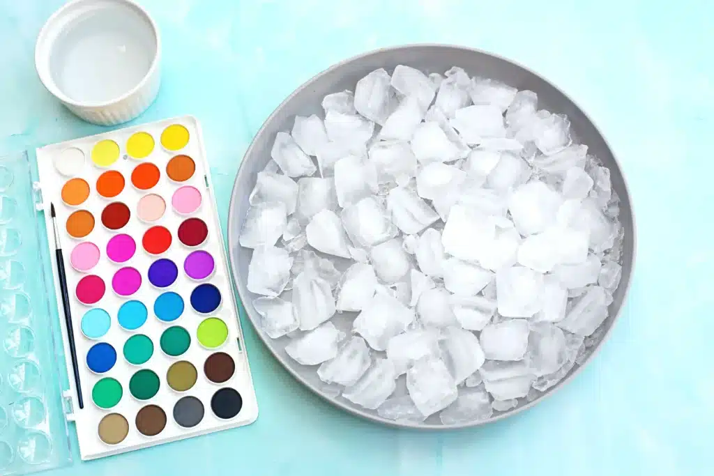 Ice Painting - Cultivating Brilliant Minds Watercolor palette with colorful round paints beside a bowl of ice on a light blue surface.