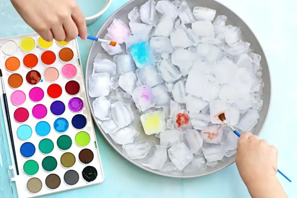 Ice Painting - Cultivating Brilliant Minds an ice painting art activity - Hands paint ice cubes in a bowl using colorful watercolors from a palette on the left.