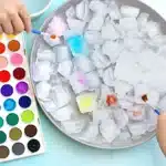 Hands paint ice cubes in a bowl using colorful watercolors from a palette on the left.