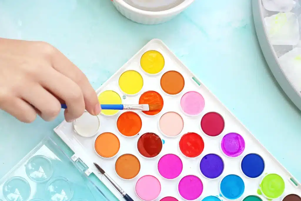 Ice Painting - Cultivating Brilliant Minds Hand holding a paintbrush over a triangular watercolor palette with bright paints on a mint surface.