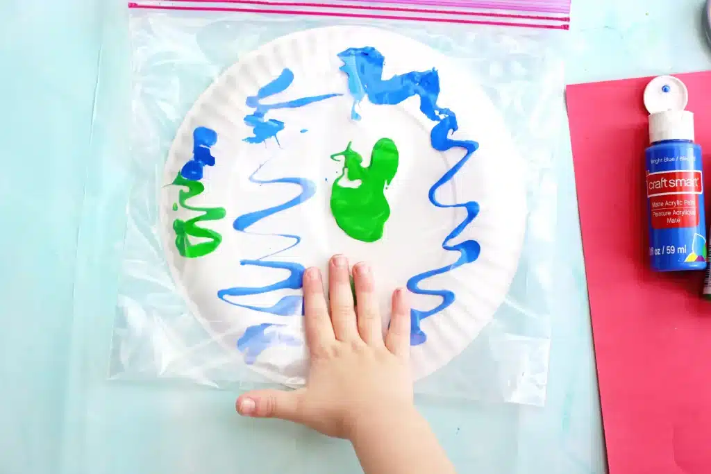 No-Mess Earth Day Painting - Cultivating Brilliant Minds a kid smooshing green and blue paint on a plate inside a plastic bag