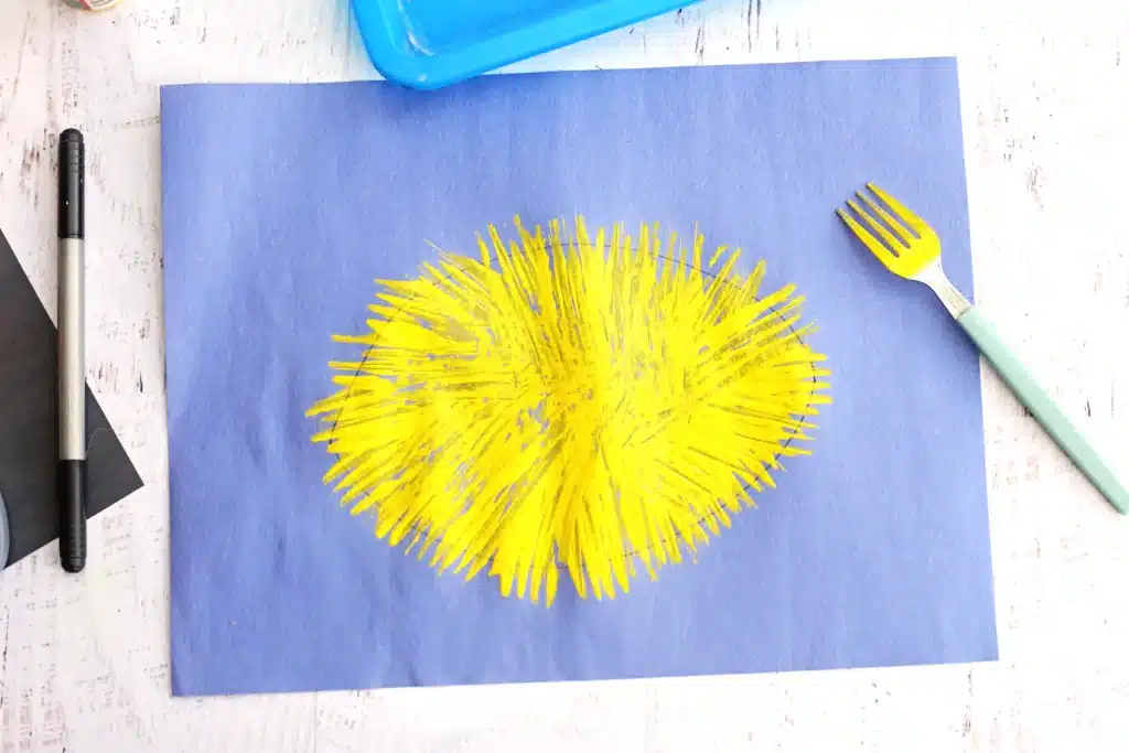Fork Painted Bee - Cultivating Brilliant Minds Blue sheet with a bright yellow spiky circular paint burst; craft tools nearby on a pale table (art desk).