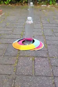 Two plastic bottles balanced upright on a rainbow-colored circular paper disc on a brick walkway.