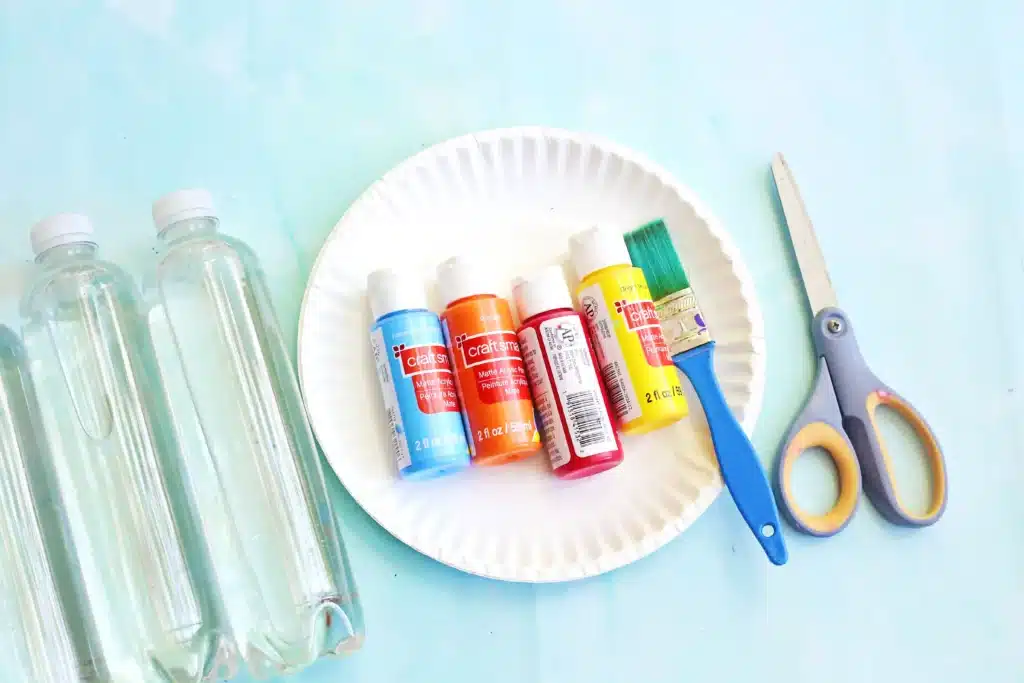 Paper Plate Ring Toss Game - Cultivating Brilliant Minds Assorted acrylic paint bottles on a white paper plate with a blue-handled brush and scissors, set beside two plastic water bottles on a light blue surface.