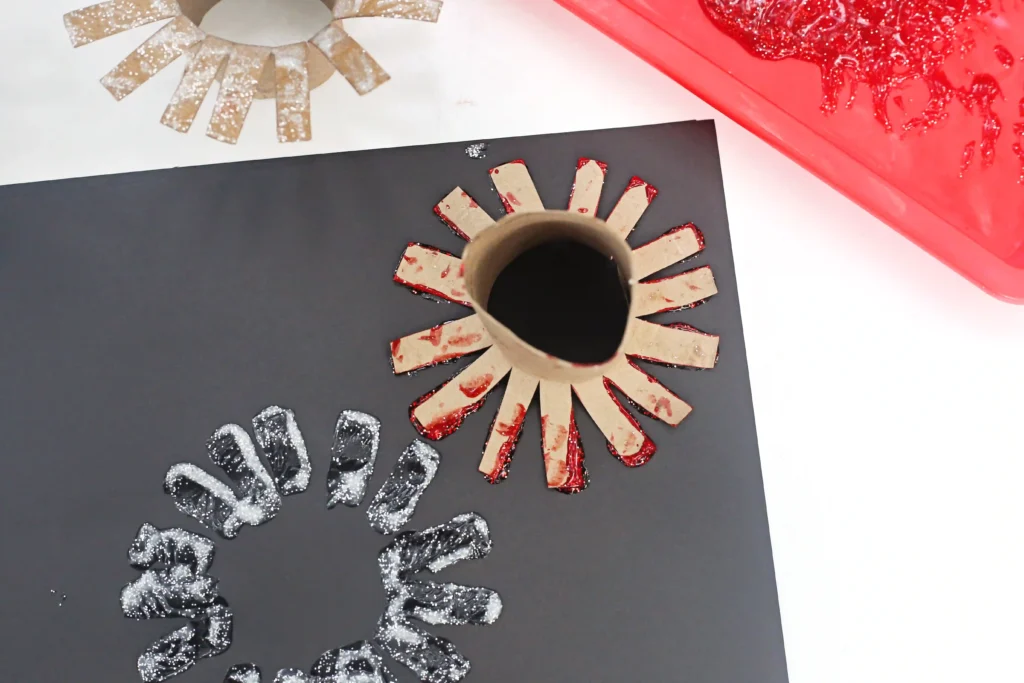 Toilet Paper Roll Firework Craft - Cultivating Brilliant Minds a circular toilet paper roll that is cut and covered with red glittery rectangular strips radiating like sun rays.