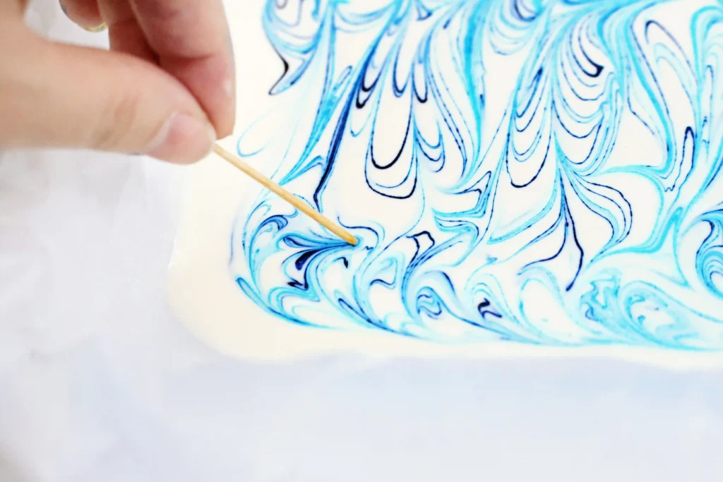 Shark Bark - Cultivating Brilliant Minds Close-up of a hand stirring blue marbled food coloring with a wooden stick on white chocolate, creating swirling patterns
