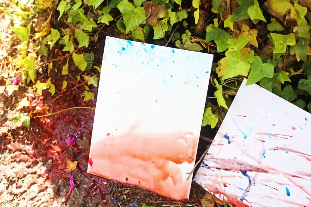 Water Balloon Splatter Art - Cultivating Brilliant Minds Two white canvases with blue, pink, and orange paint splatters leaning on ivy-covered ground outdoors.