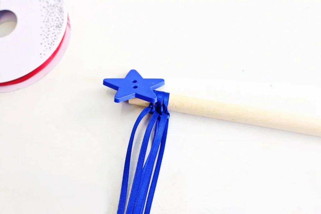 American Flag Wands - Cultivating Brilliant Minds Blue star-shaped button with blue ribbons tied to a light wooden stick on a white surface.